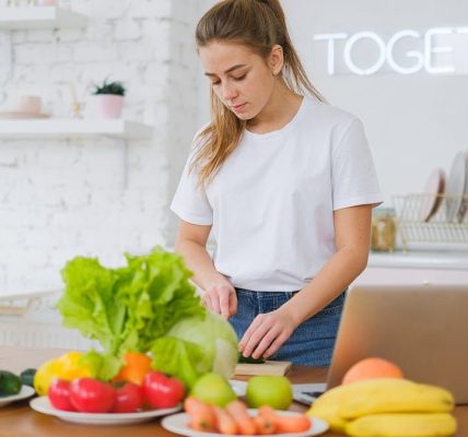 Woman in white t-shirt preparing fresh vegetables in a bright kitchen with a laptop.